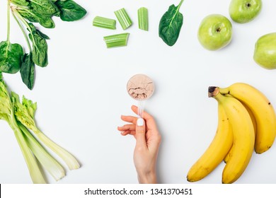 Overhead View Woman's Hand Holding Vegan Protein Powder In Scoop Near Fresh Raw Fruits And Vegetables On White Background, Flat Lay.