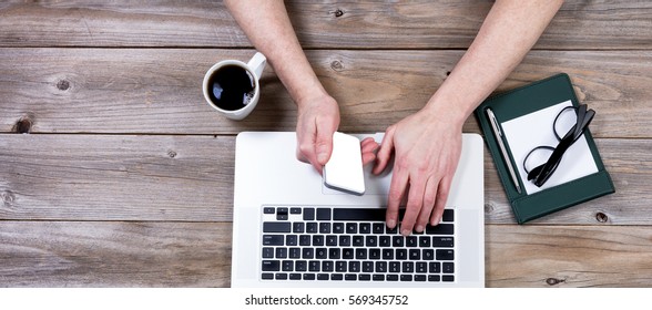 Overhead view of working desktop consisting of laptop computer, hands, coffee, cell phone, paper, pen and reading glasses