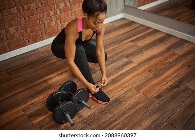 Overhead View Of A Young African Female Athlete, Attractive Woman With Perfect Muscular Body Tying Shoelaces On Her Sneakers, Next To Dumbbells Lying Down On The Floor Of A Gym