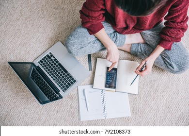 Overhead view of young brunette woman using laptop managing domestic budget alone, making notes with pencil, sitting on the floor with open laptop, documents, calculator. Businesswoman working.