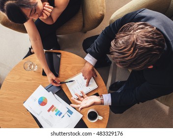 Overhead View Of Young Businessman And Woman Looking At Notepad While Sitting At Office Lobby. Coworkers Discussing New Business Project.