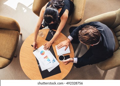 Overhead View Of Young Businessman And Woman Working On Graphs While Sitting At Office Lobby. Corporate People Discussing New Business Project Using Charts.