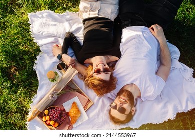 Overhead View Of A Young Couple Lying On A Picnic Blanket On A Spring Grassfield In A Countryside. Wearing Sunglasses, Smiling.