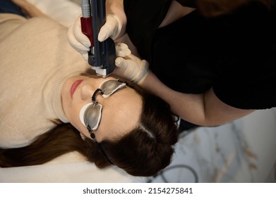 Overhead view of a young pretty European woman wearing UV protective goggles, lying down on daybed while receiving hair removal laser procedure on her face in a wellness spa clinic.