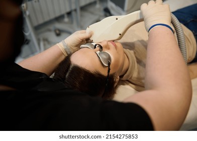 Overhead view of a young pretty European woman wearing UV protective goggles, lying down on daybed while receiving hair removal laser procedure on her face in a wellness spa clinic