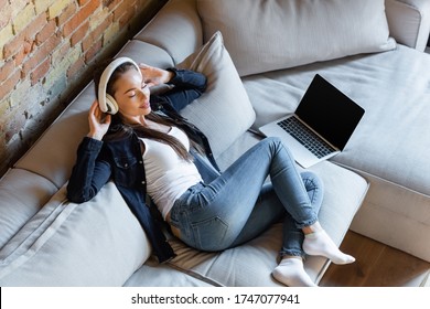 Overhead View Of Young Woman With Closed Eyes Listening Music In Wireless Headphones Near Laptop With Blank Screen