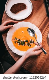 Overhead Woman's Hands Holding Spoon With Pumpkin Cream Soup, White Elegant Plate, Wooden Table. Food Photo, Food Blogger Flatlay Concept, Vertical, Toned