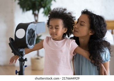 Overheated African woman and daughter turn their faces to blowing fan, electric ventilator breath fresh air in living room without air conditioning system. High temperature inside, summer day concept - Powered by Shutterstock