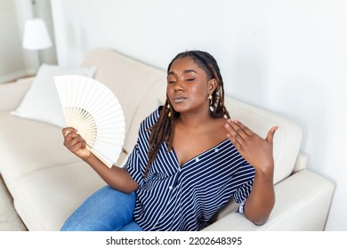 Overheated African Woman Sit On Couch At Home Feel Warm Waving With Hand Fan Cooling Down, Sweating Girl Relax On Sofa In Living Room Hold Waver Suffer From Heat, No Air Conditioner System