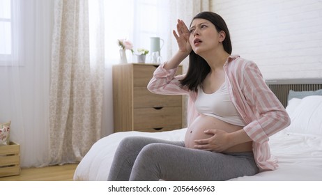 Overheated Expectant Woman Is Wiping Sweat From Her Forehead And Fanning With Hands And Shirt In Bad Mood On The Bed At Home.