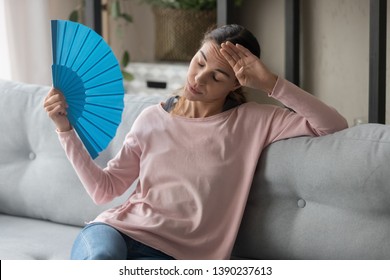 Overheated female sitting on couch in living room at hot summer weather day feeling discomfort suffers from heat waving blue fan to cool herself, girl sweating dwelling without air conditioner concept - Shutterstock ID 1390237613