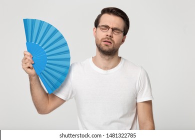 Overheated Millennial Unhappy Man In Eyewear Waving Fan, Suffering From High Temperature Inside Or Hot Summer Weather, Isolated On Grey White Studio Background. Young Guy Trying Cool Down, Sweating.