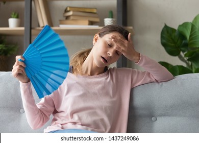 Overheated millennial woman sit on couch at home feel warm waving with hand fan cooling down, sweating girl relax on sofa in living room hold waver suffer from heat, no air conditioner system