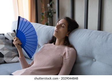 Overheated tired young Latina woman sit lean on sofa at home wave hand fan reduce heat, suffer from hot weather, lack of air conditioner indoor feels unwell exhausted, have hormonal imbalance concept