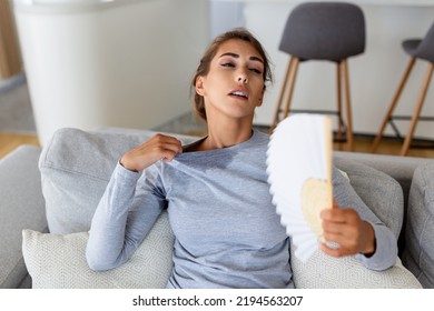 Overheated Woman Sit On Couch At Home Feel Warm Waving With Hand Fan Cooling Down, Sweating Girl Relax On Sofa In Living Room Hold Waver Suffer From Heat, No Air Conditioner System