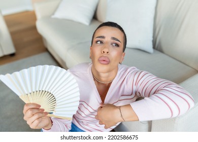 Overheated Woman Sit On Couch At Home Feel Warm Waving With Hand Fan Cooling Down, Sweating Girl Relax On Sofa In Living Room Hold Waver Suffer From Heat, No Air Conditioner System