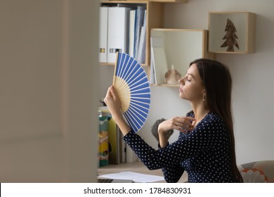 Overheated Young Caucasian Woman Sit At Desk At Home Wave With Hand Fan Suffer From Hot Weather No Air Conditioner, Sick Unwell Millennial Female Use Waver, Struggle With Hormonal Imbalance