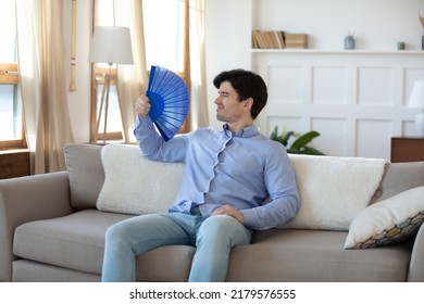 Overheated Young Man Owner Renter Tenant Of House Flat Resting On Sofa In Living Room Saving Himself From Heat Using Paper Weaver Understanding Problem Of Bad Ventilation And Lack Of Air Conditioning.