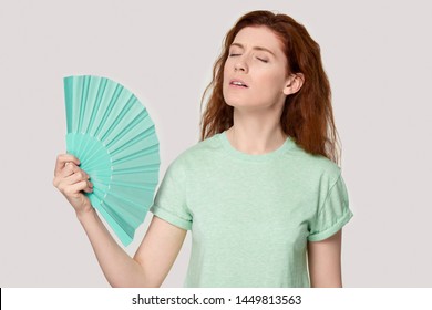 Overheated Young Red-haired Woman Using Fan, Cooling Herself, Suffering From Hot Summer Weather Or High Temperature Inside, Heat Stroke, Feeling Unwell, Sweating, Isolated On Grey Studio Background.