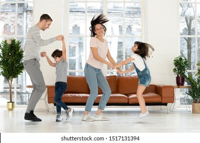 Overjoyed active parents dancing to favorite music with happy kids at living room at home. Happy dad and mom holding hands of adorable school children, twisting, having fun, celebrating occasion.