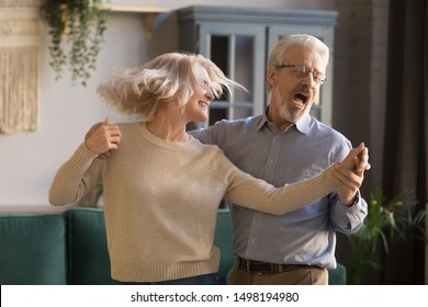 Overjoyed Active Retired Mature Couple Dancing Together Enjoy Leisure Activity In Living Room, Happy Cheerful Senior Grey-haired Husband And Wife Feel Excited Have Fun Relaxing On Weekend At Home