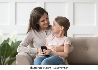 Overjoyed Adorable Daughter Holds Smart Phone Sitting On Mothers Lap Looking At Each Other Feels Happy. Modern Tech Usage, Bad Habit Kids And Electronic Gadgets, Parental Control Online Safety Concept