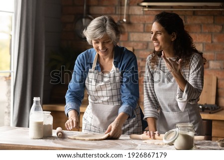 Overjoyed adult Hispanic girl with senior mom make dough bake cookies pastries together on weekend. Happy millennial Latino woman with mature mother have fun cooking dessert at home kitchen.