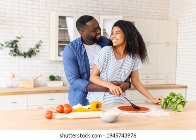 Overjoyed African American Couple In Love Standing Next To Kitchen Counter, Hugging, Spending Leisure Time Weekend Together At Home, Looking At Each Other, Cooking, Girl Is Spreading Sauce On Pizza