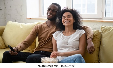 Overjoyed african American couple sit relax on cozy sofa in living room watch TV eating popcorn, happy biracial husband and wife rest on comfortable couch have fun enjoying movie leisure time at home