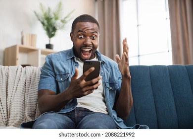 Overjoyed african american man celebrating success with yes gesture while getting some goods news on mobile. Young guy sitting on couch with sincere happiness on face.