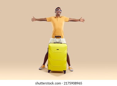 Overjoyed African American man in glasses isolated on brown studio background excited about summer vacation. Smiling biracial guy with suitcase ready for summertime travel or journey.