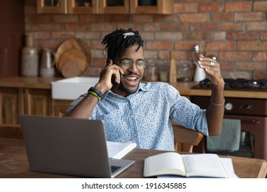 Overjoyed African American Man Talking On Phone, Distracted From Work Or Studying, Sitting At Table With Laptop At Home, Chatting With Friends During Break, Hearing Good News, Pleasant Conversation