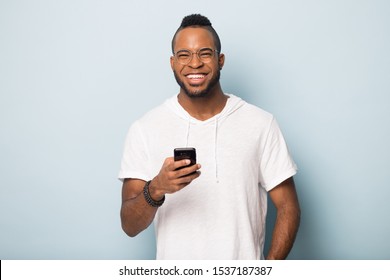 Overjoyed African American Millennial Man In Casual Wear And Glasses Isolated On Blue Studio Background Use Modern Cellphone Look At Camera, Happy Biracial Male Client Excited With New Smartphone App