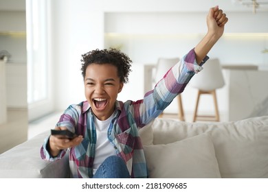 Overjoyed African American Teen Girl Celebrating Goal, Sitting On Couch In Living Room At Home. Excited Happy Young Woman Watching Tv Football Play, Rooting For Favorite Sport Team, Shouting.