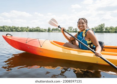 overjoyed african american woman in life vest paddling in sportive kayak and smiling at camera on lake with green picturesque shore on background in summer - Powered by Shutterstock