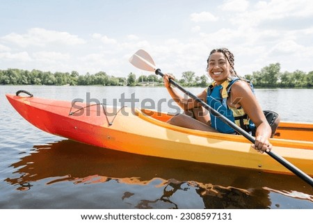 overjoyed african american woman in life vest paddling in sportive kayak and smiling at camera on lake with green picturesque shore on background in summer