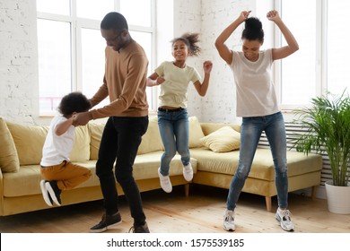 Overjoyed African American Young Family Have Fun Enjoy Leisure Weekend At Home Together, Happy Biracial Parents With Small Preschooler Kids Dance Jump Entertain In Living Room, Stress Free Concept