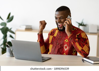 Overjoyed African Businessman In Traditional Clothes Celebrating Success At Workplace In Office, Talking On Cellphone And Looking At Laptop Screen, Emotionally Reacting To Good News, Free Space