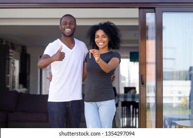 Overjoyed African Married Couple Standing On Summer Terrace Porch Near New Modern House. Happy Wife Hold Keys Embracing Beloved Husband Showing Thumbs Up Sign. Property Ownership, Loan Tenancy Concept