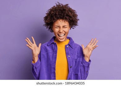 Overjoyed Afro American Girl Exclaims Loudly Keeps Hands Raised Feels Excited And Very Happy To Hear Awesome News Dressed In Velvet Jacket Isolated Over Purple Background. People And Emotions Concept