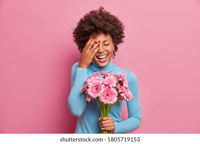 Overjoyed Afro American woman laughs out loudly, keeps palm on face, stands indoor with bouquet of gerberas, going to congratulate mother with womens day, receives flowers from unknown person - Powered by Shutterstock