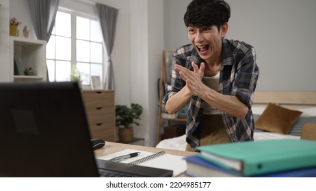 Overjoyed Asian Chinese Male Student Using Phone At Background Is Rushing To Check Email And Screaming With Fist For His Success While Typing On Laptop