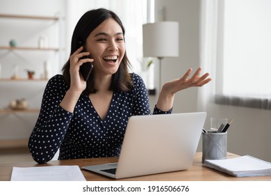 Overjoyed Asian Female Laughing Telling Funny Story By Phone To Friend Colleague Sitting By Office Computer Screen. Young Vietnamese Woman Enjoying Conversation By Cell Discussing Good Positive News