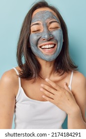 Overjoyed Asian Woman Has Dry Skin Applies Nourishing Clay Mask For Moisturising Smiles Broadly Keeps Hand On Chest Tries To Improve Symptoms Of Dermatitis Wears White T Shirt Isolated On Blue Wall