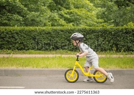 Overjoyed baby boy in safety helmet speed riding on balance bike at summer park green trees enjoy happy childhood. Playful male kid fast running driving cycle sports leisure activity side view