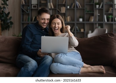 Overjoyed Beautiful Millennial Couple Sit Relax On Cozy Couch Look At Laptop Screen Read Great News, Got Fantastic Offer, Winning Lottery Online, Celebrate Moment Of Auction Victory. Good News Concept