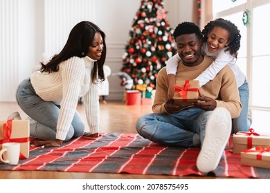 Overjoyed Black Family Exhanging Christmas Presents, Happy Dad Unpacking Opening Wrapped Gift Box From Daughter And Wife, People Sitting On Floor Blanket Near Decorated Tree At Home In Living Room