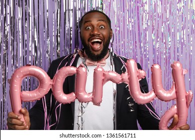 Overjoyed black gentleman with bristle, dressed in formal suit, entertains guests, holds party accessories, poses in photozone against purple background with sparkled long tinsel. Holiday celebration