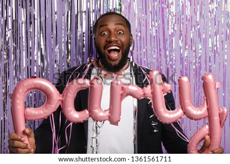Overjoyed black gentleman with bristle, dressed in formal suit, entertains guests, holds party accessories, poses in photozone against purple background with sparkled long tinsel. Holiday celebration