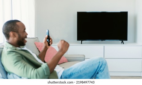 Overjoyed Black Man Watching Sports Game On TV, Celebrating Victory And Shaking Fists, Sitting On Sofa, Looking At Empty Screen, Mockup, Selective Focus